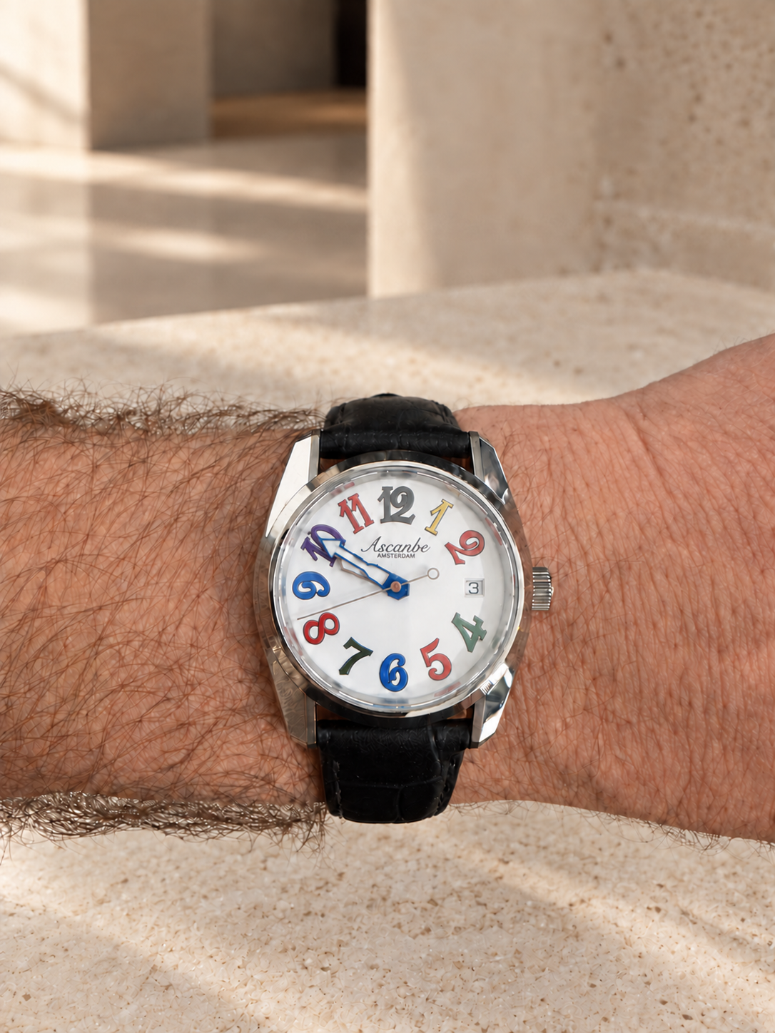 A one-off Ascanbe watch with multicoloured numerals on a black leather strap, worn on the wrist